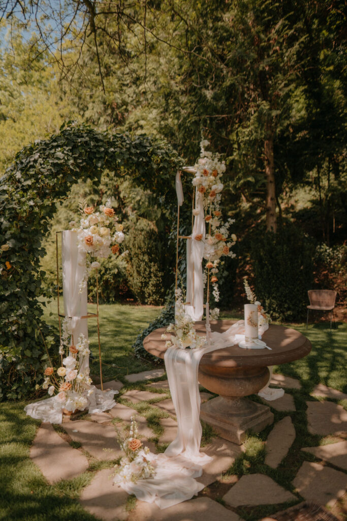 Florale Hochzeitsdekoration mit luftigem Stoff, rustikalem Trautisch und Blüten in Pastell und Orange im Schlossgarten