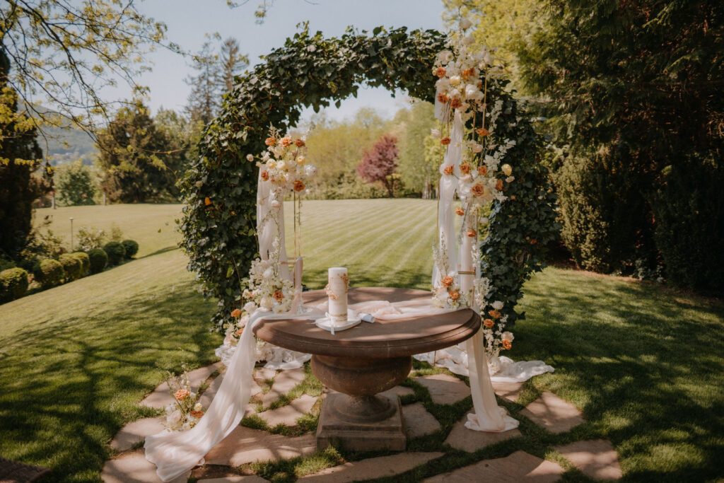 Romantisch dekorierter Trautisch unter grünem Efeubogen mit floraler Hochzeitssäule in Creme und Rostorange im Garten von Aiola im Schloss St. Veit