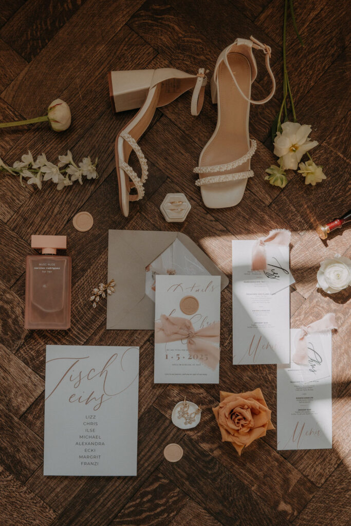 Hochzeitseinladung, Papeterie, Brautschuhe und Accessoires elegant arrangiert auf Holzboden – Hochzeit von Lizz & Chris im Aiola im Schloss St. Veit, fotografiert von Pixellicious.