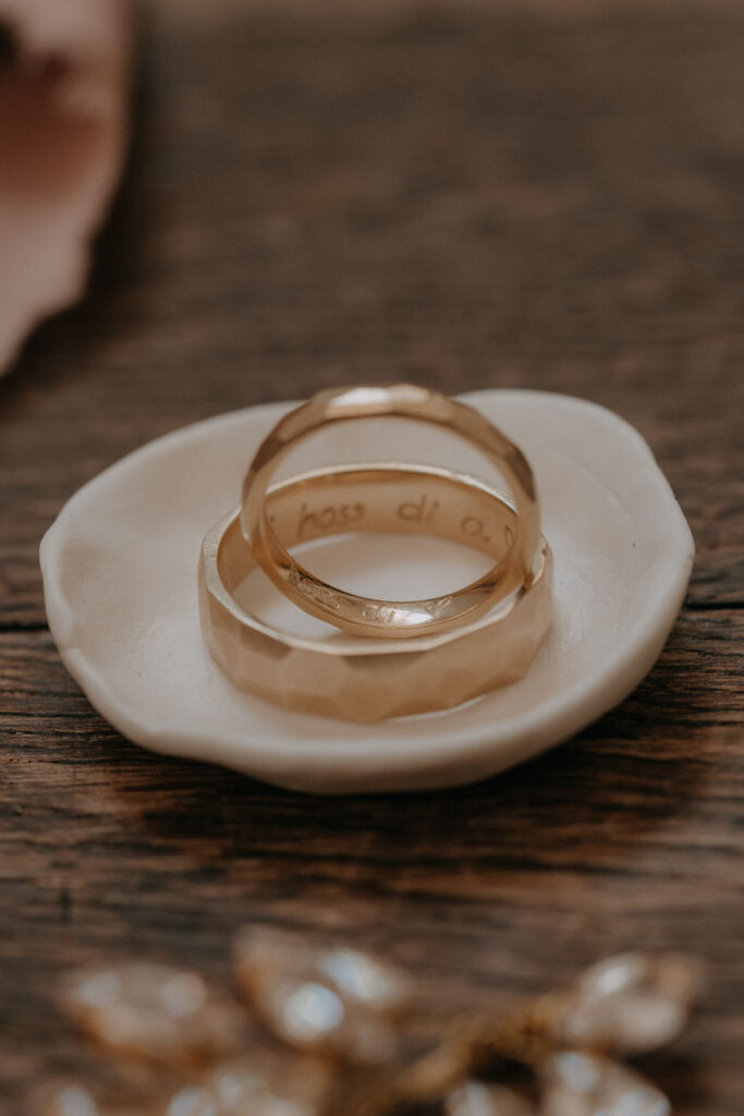 Goldene Eheringe mit Gravur „hass di“ in einer Keramikschale – Detailaufnahme der Hochzeit von Lizz & Chris in der Steiermark.