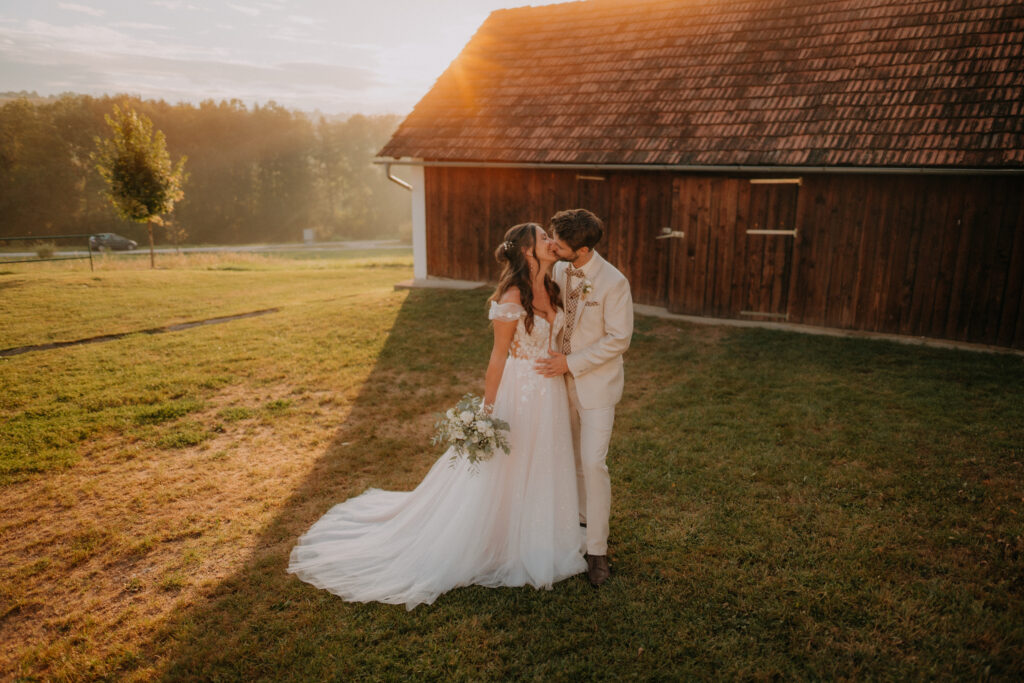 Brautpaar küsst sich bei Sonnenuntergang vor Holzstadl – natürliche Hochzeitsfotografie