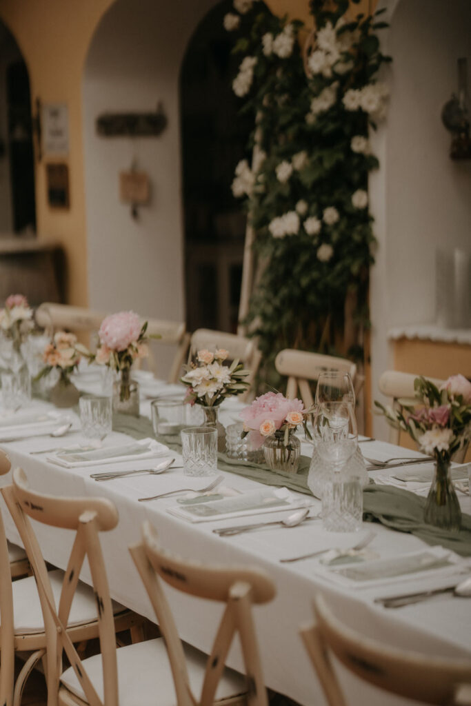 Lange Tafel im Freien, elegant gedeckt mit weißen Stühlen, Glasvasen mit rosa Blumen und grünem Tischläufer, perfekte Sommerhochzeit.