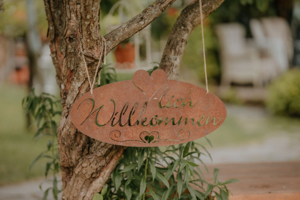 Rustikales Holzschild mit der Aufschrift „Herzlich Willkommen“ in Herzform, umgeben von grünen Pflanzen.