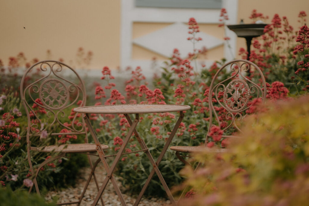 Rustikaler Gartenstuhl und Tisch mit roten Blüten im Hintergrund, der Charme des Gartens bei der Hochzeitslocation.
