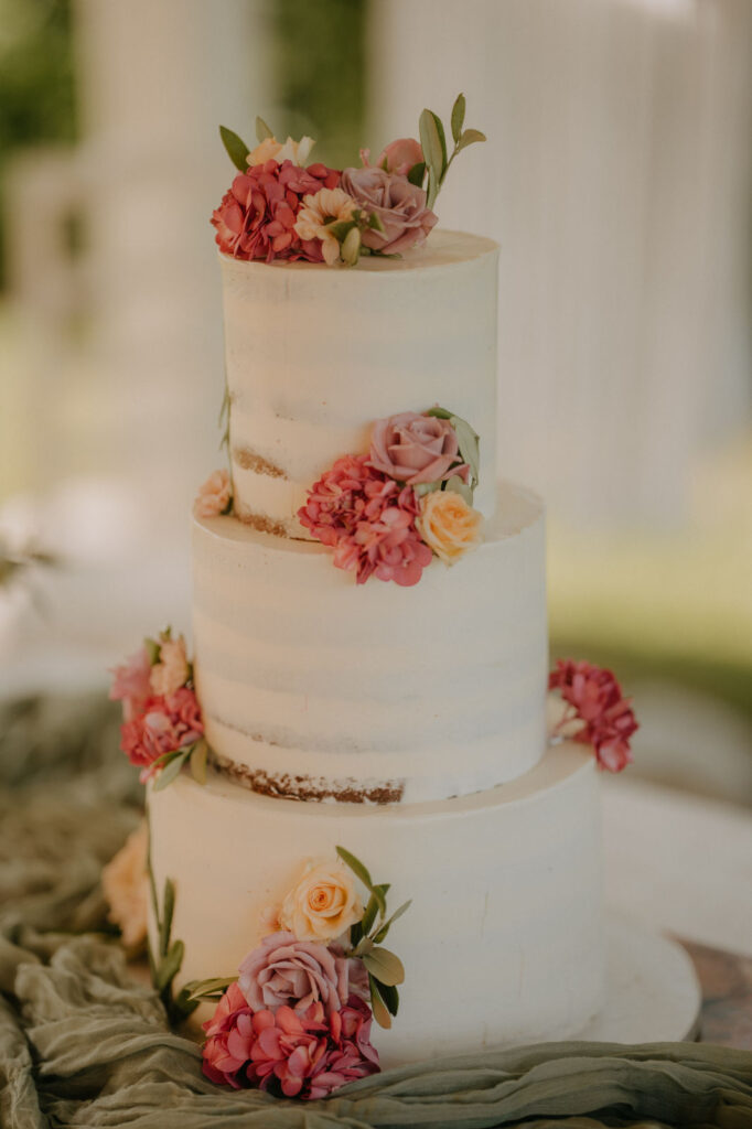 Elegante dreistöckige Hochzeitstorte, dekoriert mit zarten rosa Blüten, vor dem warmen Licht des Gartens.