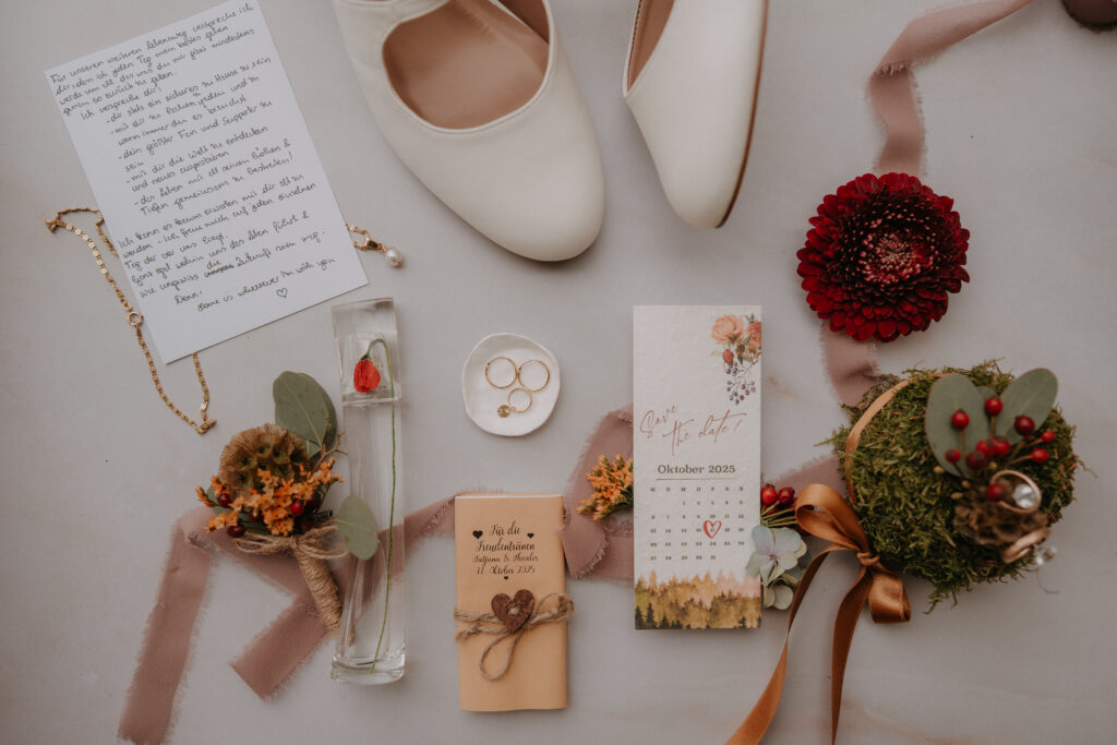 Flatlay mit Hochzeitsschuhen, Ringen, floralen Accessoires, handgeschriebenem Brief, Save-the-Date-Karte und herbstlichen Details.