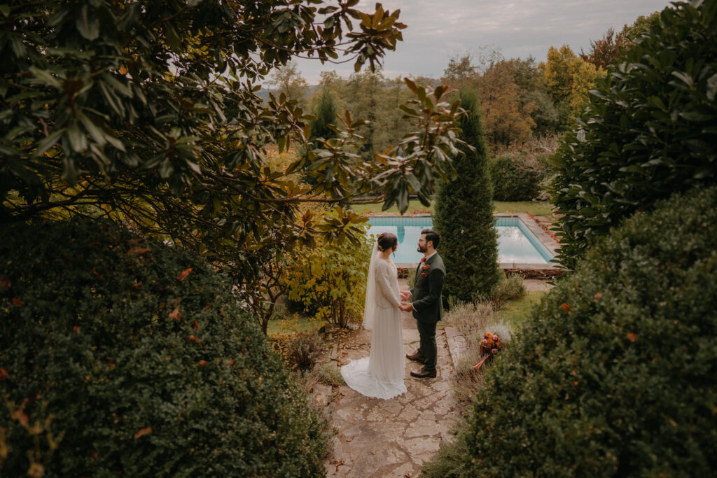 Braut und Bräutigam stehen sich auf einem Gartenweg gegenüber, eingerahmt von Sträuchern, mit Blick auf den Pool.