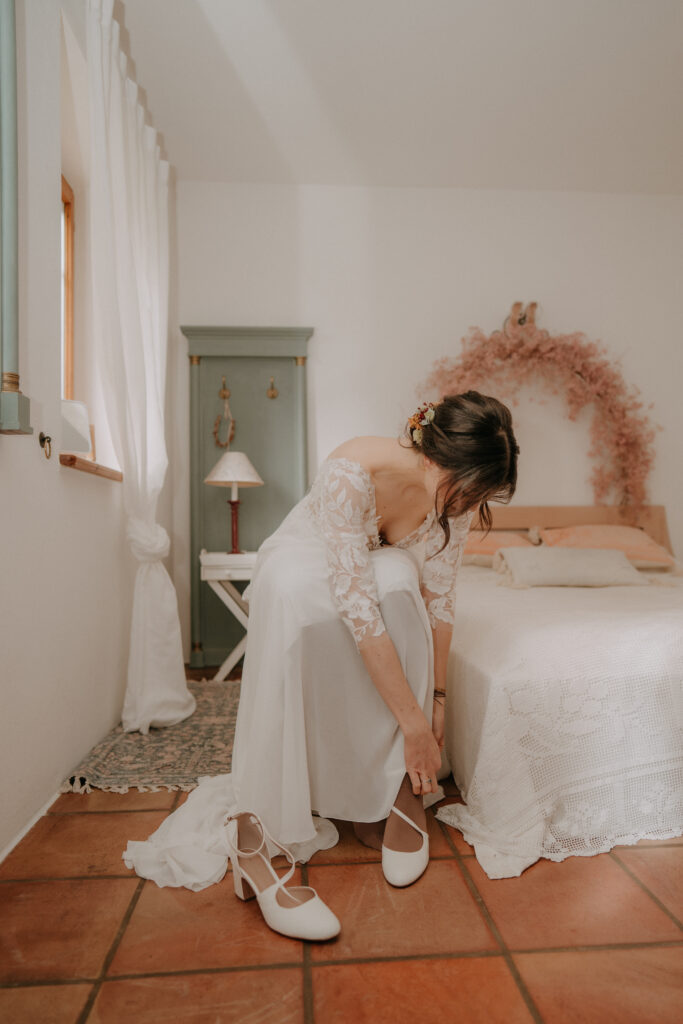 Die Braut sitzt im hellen Zimmer und zieht ihre weißen Brautschuhe an, während ihr langes Kleid locker über den Boden fällt.