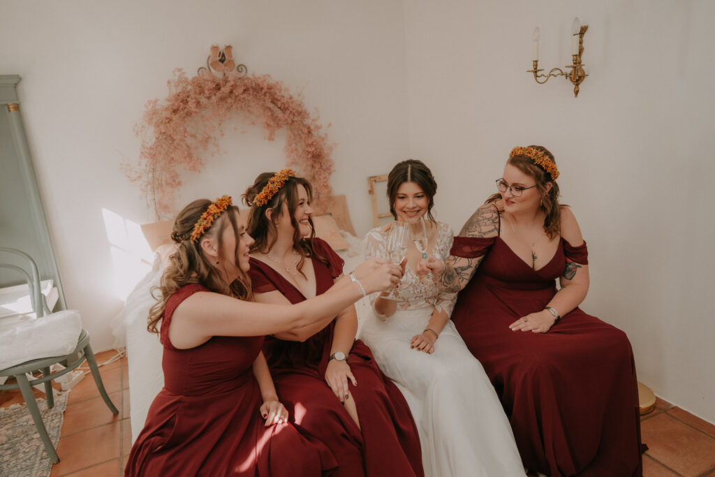 Die Braut sitzt mit ihren Brautjungfern in bordeauxroten Kleidern auf dem Bett und stößt beim Getting Ready mit Sektgläsern an.