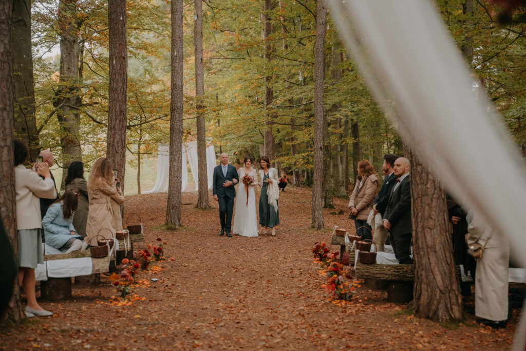 Die Braut wird im herbstlich geschmückten Wald von ihren Eltern zum Traubogen geführt, während die Gäste aufstehen und den Moment festhalten.