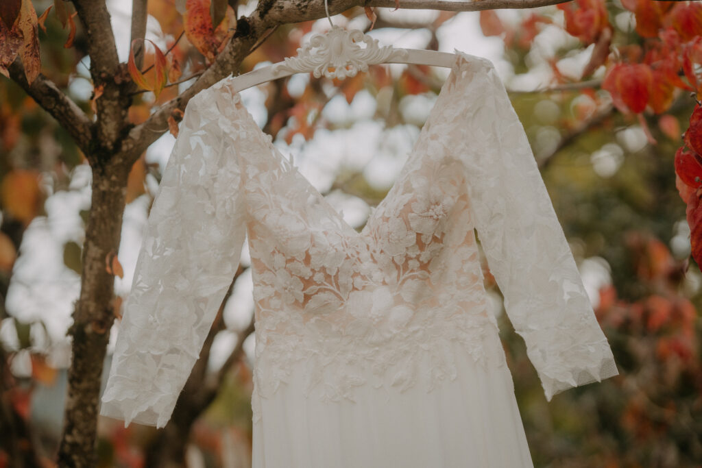 Spitzen-Brautkleid mit langen Ärmeln hängt an einem Ast zwischen bunten Herbstblättern im Garten.