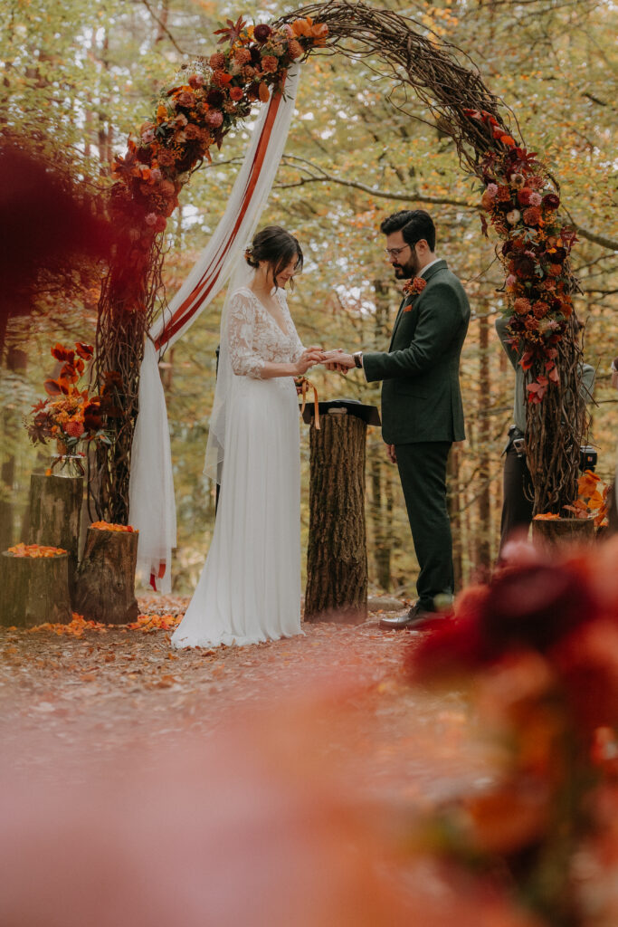Braut und Bräutigam tauschen ihre Ringe unter einem herbstlich dekorierten Traubogen im Wald.