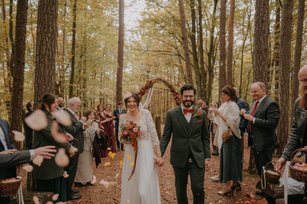 Braut und Bräutigam gehen lächelnd durch ein Spalier aus Familie und Freunden bei ihrer herbstlichen Waldhochzeit, während Blütenblätter geworfen werden.