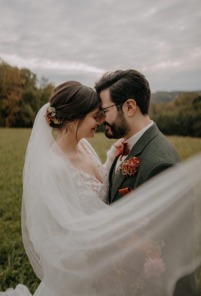 Nahaufnahme des Brautpaares, Stirn an Stirn, umgeben vom weichen Schleier im Freien.