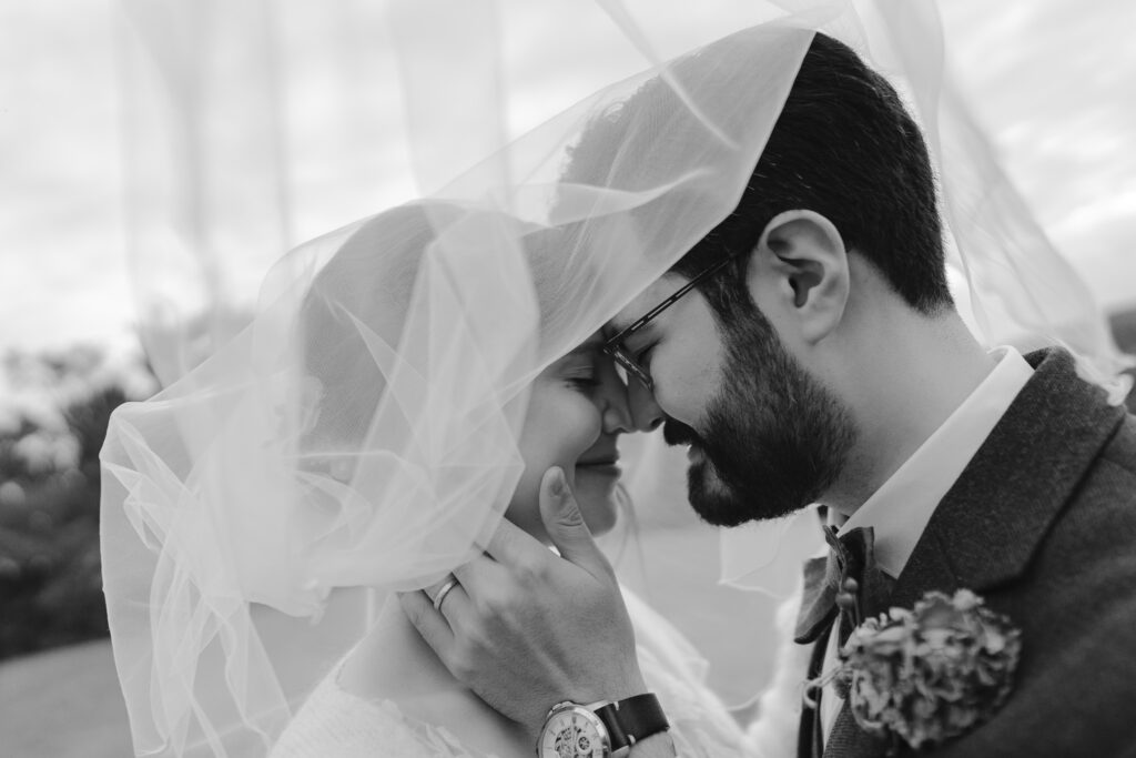 Braut und Bräutigam stehen Stirn an Stirn unter dem Schleier, sanfter und emotionaler Moment in Schwarz-Weiß.