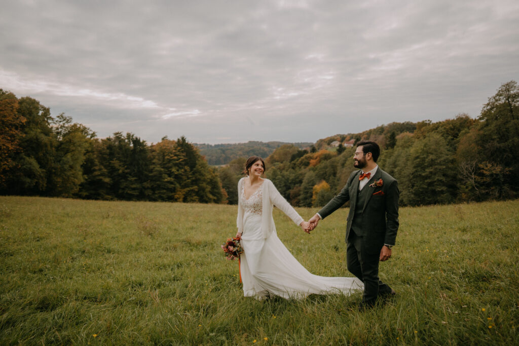 Braut und Bräutigam gehen Hand in Hand über eine weite Wiese, lächelnd vor herbstlicher Landschaft.