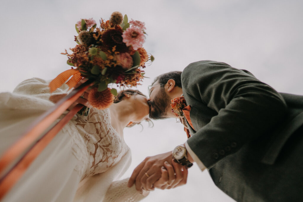 Braut und Bräutigam küssen sich aus der Froschperspektive, der herbstliche Blumenstrauß im Vordergrund.
