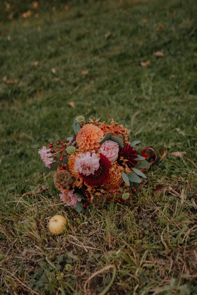 Herbstlicher Brautstrauß mit Dahlien, Beeren und Rosen in warmen Rot- und Orangetönen, liegend auf einer Wiese bei einer Waldhochzeit in der Steiermark.