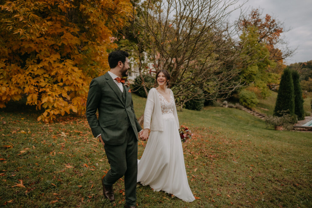 Braut und Bräutigam gehen Hand in Hand über eine Wiese mit leuchtend gelbem Herbstlaub.
