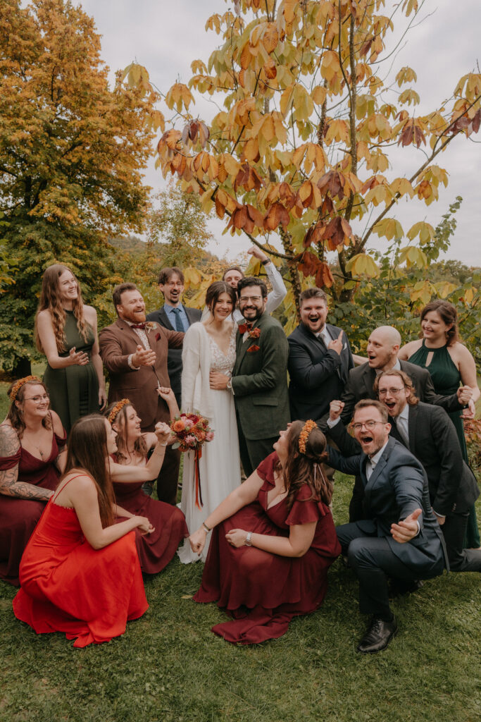 Große fröhliche Gruppe rund um das Brautpaar, alle jubeln inmitten bunter Herbstbäume.