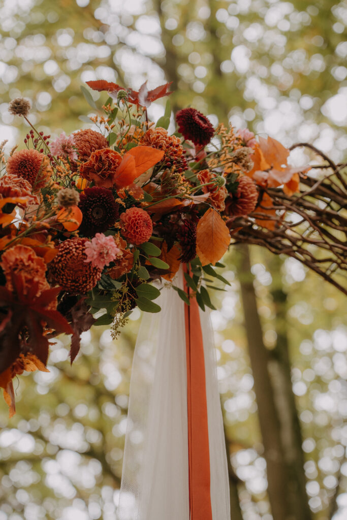 Nahaufnahme des herbstlich dekorierten Traubogens mit orangefarbenen Blüten, Zweigen und Stoffbändern im Wald.