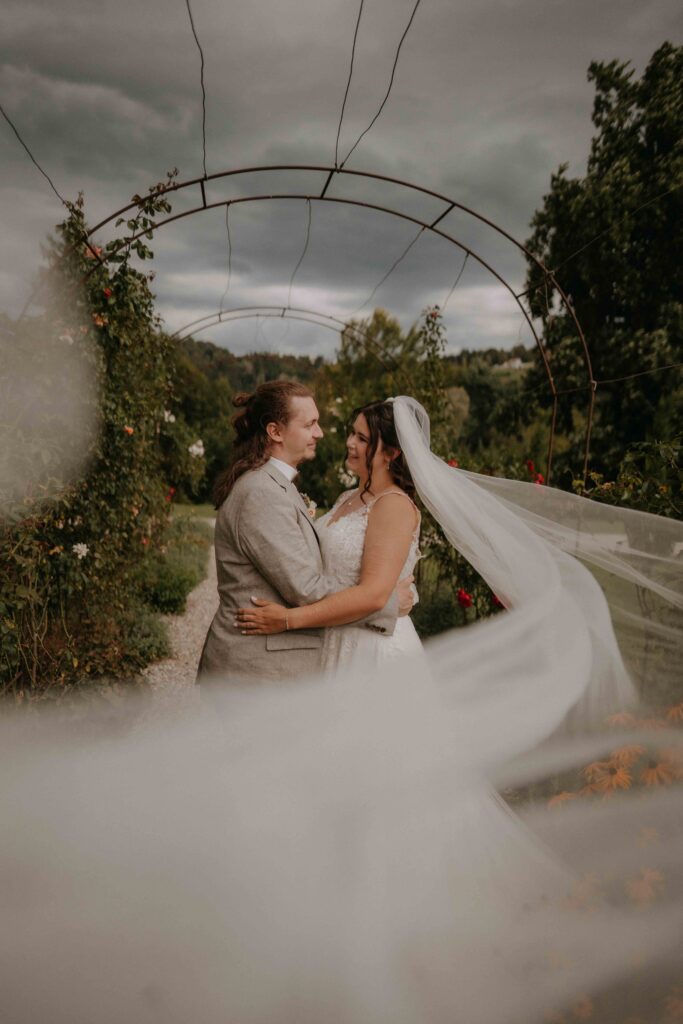 Brautpaar im romantischen Bodennebel unter einem Rosentor im Schloss Ottersbach – Hochzeitsfotografie Steiermark
