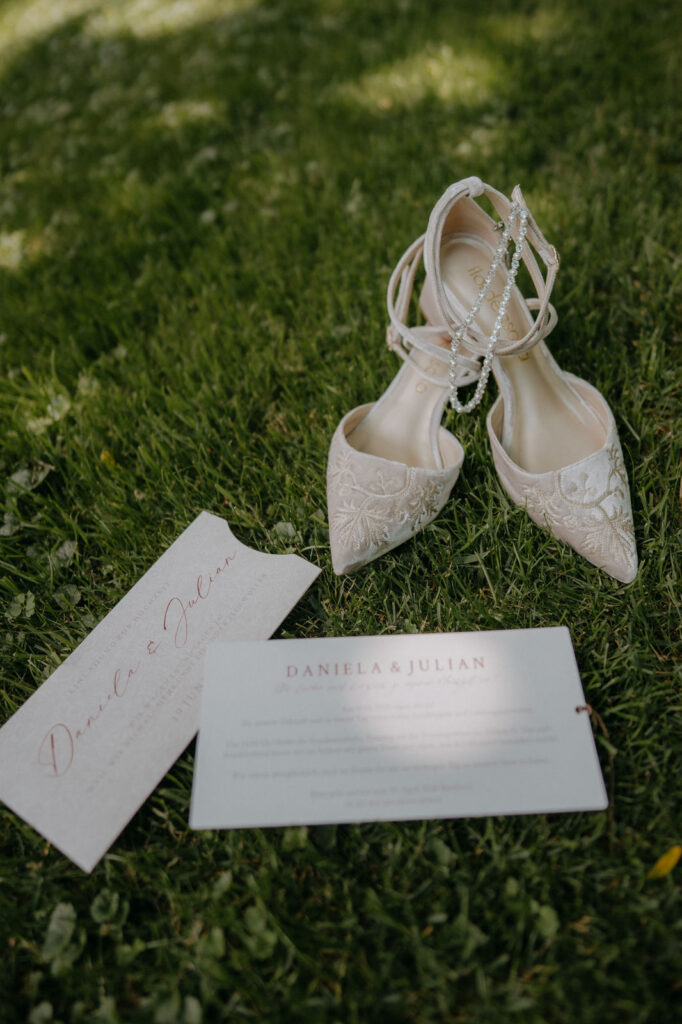 Brautschuhe mit Spitze und Einladungskarte auf Rasen – Hochzeit Aiola im Schloss Graz