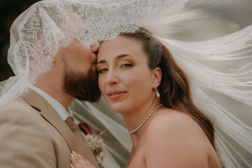 Bräutigam küsst Braut auf Stirn durch Schleier – Hochzeit Aiola im Schloss Graz