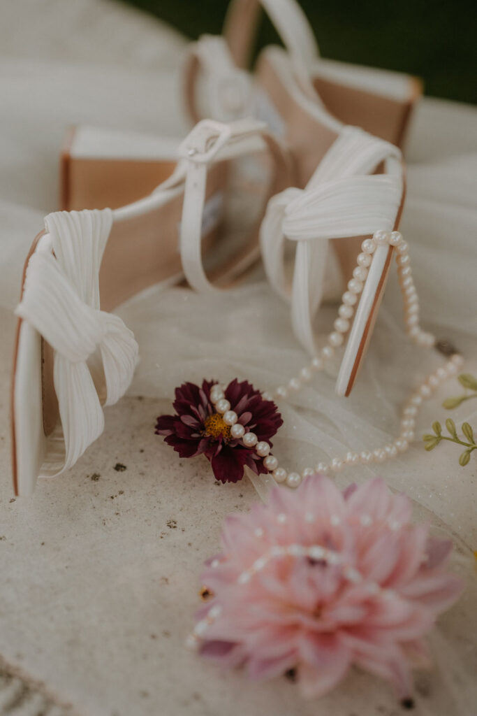 Weiße Brautschuhe mit Perlenkette und Dahlien auf Tüll drapiert, Detailfoto von der Hochzeit auf Schloss Ottersbach