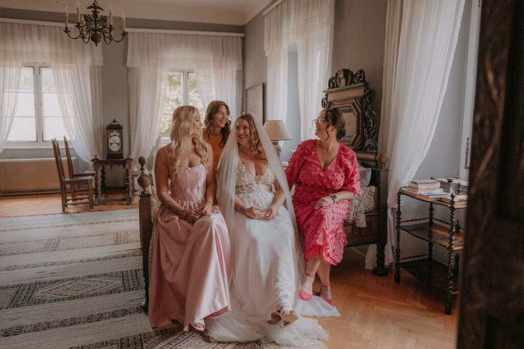 Braut sitzt lachend mit ihren drei Brautjungfern in Rosa und Pink vor der Trauung auf Schloss Ottersbach