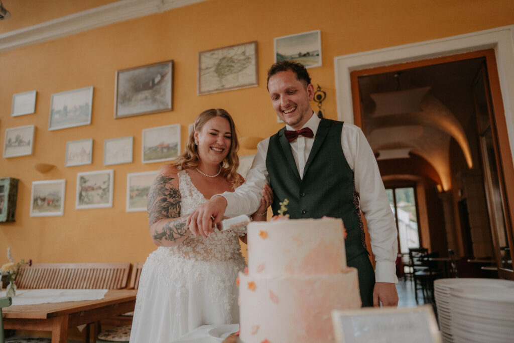 Brautpaar schneidet lachend die dreistöckige Hochzeitstorte an, im Hintergrund gerahmte Landkarten an oranger Wand auf Schloss Ottersbach