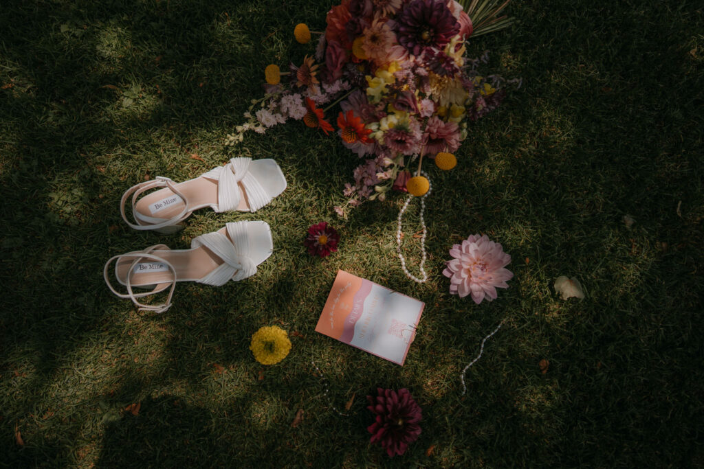Flatlay mit weißen Brautschuhen, buntem Blumenstrauß und Einladungskarte auf grüner Wiese bei der Hochzeit auf Schloss Ottersbach