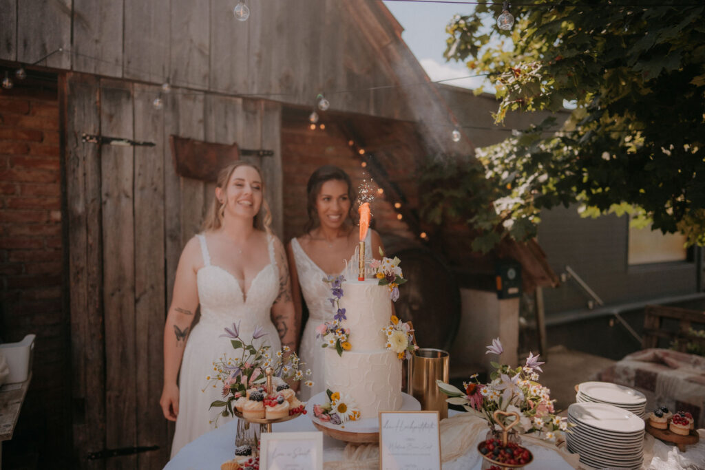 Bräute stehen lächelnd neben der Hochzeitstorte mit Wunderkerze