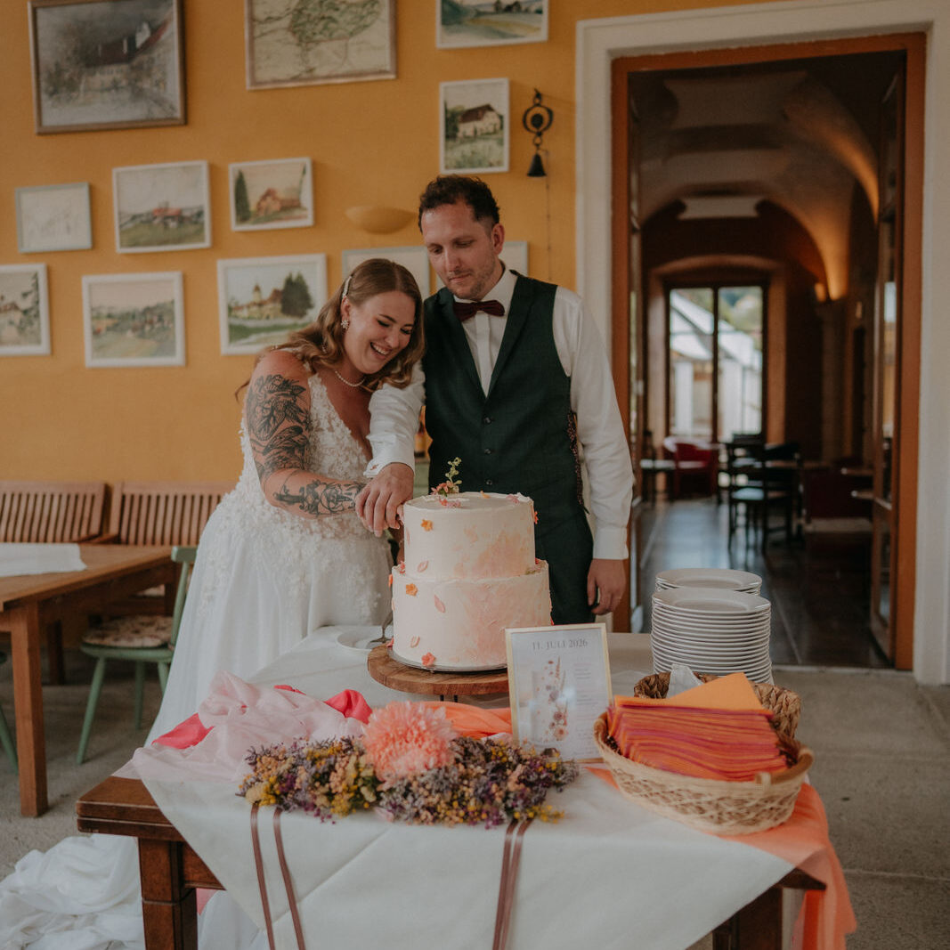 Brautpaar schneidet lachend die Hochzeitstorte an vor gerahmten Landkarten auf Schloss Ottersbach