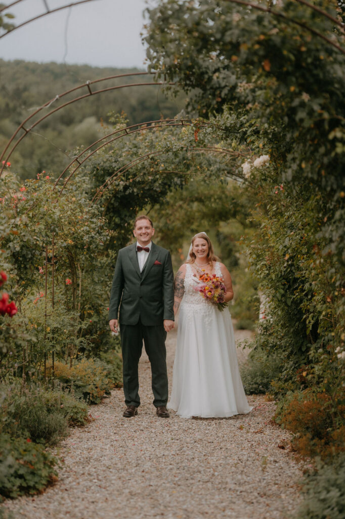 Brautpaar steht Hand in Hand unter einem grünen Rosenbogen auf dem Kiesweg von Schloss Ottersbach